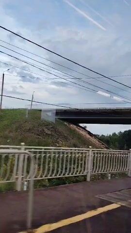 The video shows a view from inside a moving train, looking out at a train station platform. The platform has a metal fence, a yellow line on the ground, and a covered area with a metal roof supported by pillars. There are also streetlights and power lines visible outside the station. In the background, there is a grassy embankment with trees and a bridge crossing over the tracks. The sky is cloudy.