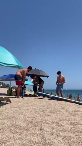 A group of people are on a sandy beach on a sunny day. Two individuals, a man and a woman, are standing under a black umbrella, seemingly interacting with a blue object on the ground. The man is wearing red and blue swim trunks, and the woman is wearing a black swimsuit. To their left, a man in red and blue swim trunks is leaning over, also near the blue object. To their right, a man in blue swim trunks stands, facing away from the camera. In the background, there are more beach umbrellas, people swimming and relaxing in the ocean, and a clear blue sky. A long, thin object, possibly a paddleboard or surfboard, is lying on the sand in the foreground.