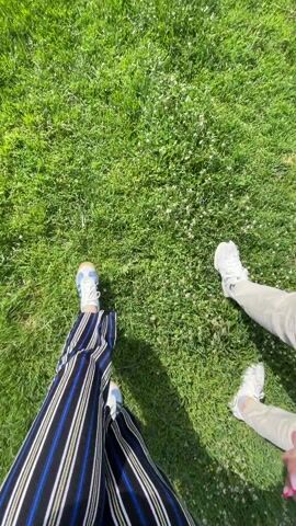 A first-person view shows two individuals standing on grass. The person filming is wearing striped pants, with lines of blue, black, white, and yellow. The individual is also wearing tennis shoes with blue and white coloring. To the right, the other person is wearing white shoes and beige pants. The green grass has some small, white flowers.