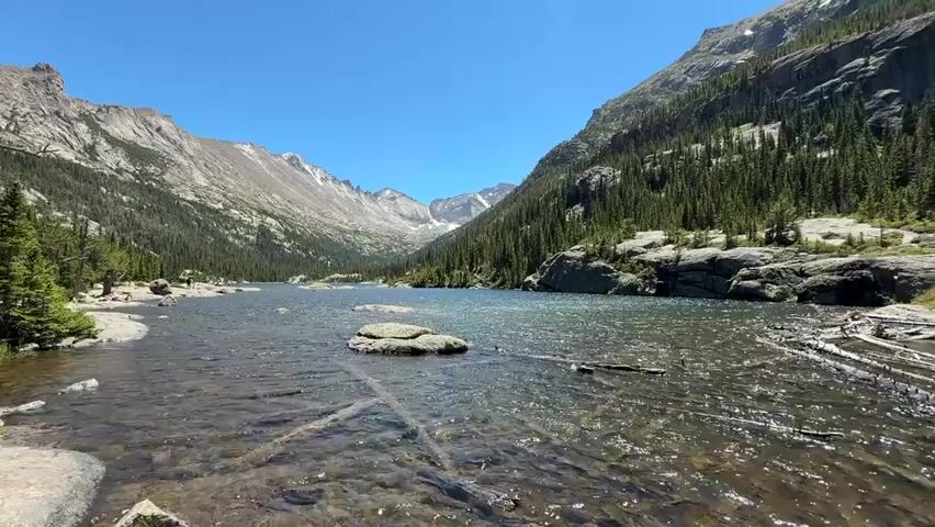 This is a first-person view video of a person walking along the edge of a clear lake. The lake is surrounded by mountains covered in evergreen trees. The sky is clear and blue. The water is shallow near the shore, and there are rocks and fallen logs visible. The person is wearing dark clothing and walking to the right of the frame. In the distance, a person in a light-colored shirt can be seen standing near the water. The scene appears to be a natural landscape, possibly a national park or wilderness area. The video is shot in landscape mode and appears to be on a sunny day.