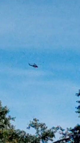 THE VIDEO SHOWS A HELICOPTER IN THE SKY ABOVE TREES AND A ROOF. THE SKY IS BLUE AND CLEAR. THE TREES ARE GREEN AND LUSH. THE ROOF IS DARK COLORED AND TILED. THE HELICOPTER IS SMALL AND APPEARS TO BE FLYING AT A HIGH ALTITUDE. THE VIDEO IS TAKEN FROM A LOW ANGLE, LOOKING UP TOWARDS THE SKY. THE FOOTAGE IS GRAINY AND OUT OF FOCUS, MAKING IT DIFFICULT TO IDENTIFY ANY SPECIFIC DETAILS ABOUT THE HELICOPTER OR THE SURROUNDING ENVIRONMENT.