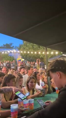 The video shows a crowded outdoor concert. People are close together, with some holding drinks and phones. A large speaker system and lights with smoke effects are visible above the crowd. The event appears to be taking place at dusk or night, with string lights illuminating the area.