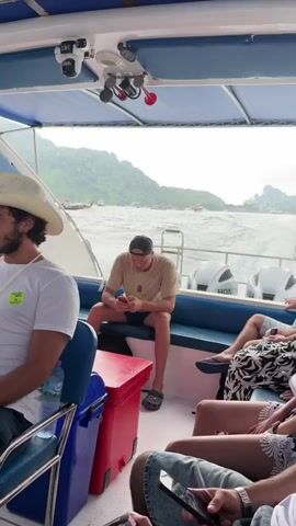 The video shows the interior of a speed boat with several passengers. The perspective is from the back of the boat, looking forward. In the foreground, a person in a white shirt and a cowboy hat is seated on the left. To their right, a person in a tan t-shirt and a baseball cap is looking at their phone. Further back, other passengers are visible, some looking out the window or at their phones. The boat is moving, and waves are visible in the water outside. In the distance, a mountainous landscape with some trees and other boats can be seen. The sky appears overcast. There are also life jackets visible on the left side of the boat. The boat has blue seating and a blue canopy.