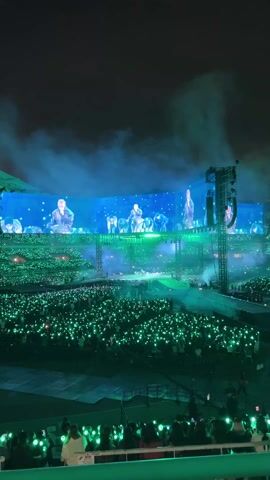 The video shows a large crowd of people holding up green lights at a concert. A massive screen behind the stage displays images of a group of men, who then appear on the stage itself. Smoke fills the air, adding to the atmosphere of the event. The scene is captured at night in a stadium setting.