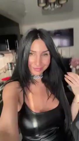 The video shows a woman indoors, possibly in her kitchen, as she is standing near a kitchen counter. She has dark, long hair and wears a black, shiny dress with thin straps and a chunky silver necklace. She smiles and looks at the camera. In the background, there is a TV on a wall, a chandelier, and various kitchen items. She holds the camera in her hand, making the video a selfie.