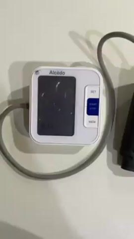 The video shows a white Alcédo brand blood pressure monitor on a white surface with a connected gray tube leading to a black cuff. The digital display is dark, but shows a faint time reading of 16:57, then 16:58. The monitor has three buttons: SET, START/STOP, and MEM. A voice states "correct level. Start measuring now."