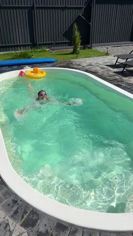 A young woman with dark hair, wearing a black bikini and swimming goggles, is treading water in a small, oval-shaped swimming pool. The pool is filled with clear, turquoise water, and the bottom is visible. Sunlight reflects off the water's surface, creating shimmering patterns. A yellow inflatable ring with a red accent is floating near the left edge of the pool. The pool is surrounded by a paved area made of dark gray and black interlocking stones. In the background, a tall, dark gray wooden fence runs along the perimeter of the property. To the left of the pool, a green lawn is visible with a small, slender tree. Two black lounge chairs are positioned on the paved area to the right of the pool. The woman is smiling and looking towards the camera. She is moving her arms as if swimming or playing in the water. The video is shot from a slightly elevated angle, looking down into the pool.