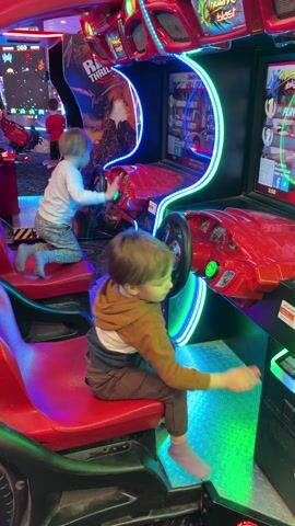 Two young children are playing a racing game at an arcade. The game consists of two bright red, car-shaped seats with steering wheels and screens, decorated with neon lights. The child in the foreground is wearing a tan hooded sweater, brown pants, and gray socks. He’s sitting in his seat and reaching to touch buttons on the game console. The other child is wearing a white shirt, gray pants, and gray socks. He is kneeling in his seat, holding the steering wheel. There’s another game machine in the background with another child playing. The arcade has colored lights on the floor and above the machines. There is a girl in a red hoodie walking in the background.