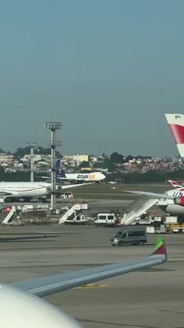 A short vertical video shows an airplane in an airport. The camera is positioned inside an airport, looking out at the tarmac through a window, with the wing of another plane in the foreground. In the midground, a British Airways plane with the tail number G-XMRA is parked, and a United Airlines plane is parked further back. An Atlas Air plane can be seen taxing in the background, along with a few airport vehicles and buildings. The sky is clear and blue.
