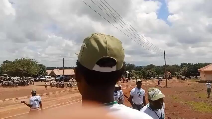 The video shows a field on a cloudy day, possibly a training ground or camp. In the foreground, people wearing white shirts and green caps, bearing the acronym "NYSC", are walking towards the camera.  The background shows a cluster of buildings with red roofs, vehicles, and groups of people gathered in the open space. An electric tower stands in the distance. The ground is mostly brown, and the sky is filled with clouds.