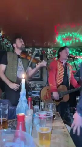 Two musicians are performing on a table in what appears to be a pub. The musician on the left is playing the violin and wearing a dark t-shirt under a military-style green vest. He has a beard and dark hair. To his right, the other musician is playing a guitar and singing into a microphone. He’s dressed in a red military jacket with gold trim.  A candle burns on the table next to the violinist, alongside several glasses of beer and some bottles. The ceiling of the pub is decorated with Christmas lights and greenery. People are gathered around the table, watching the performance.
