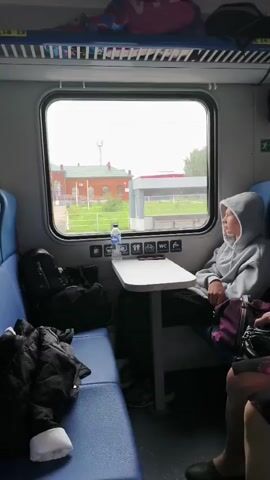 The video shows the interior of a train compartment with blue seats and overhead luggage racks. A person wearing a grey hoodie sits by the window, looking out at the passing scenery. A table is in front of them with a water bottle and other items. Luggage is visible on the seats. The scenery outside the window includes buildings, trees, and a pedestrian bridge. The train appears to be moving.
