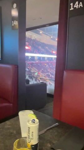 The video shows a view from a private box at a sporting event, likely a hockey game.  The box has a window overlooking the arena, with a crowd of people visible.  Inside the box, there is a table with a drink, a coaster, and a knife.  The seating in the box consists of red leather booths. A TV is mounted on the wall, showing a golf tournament leaderboard with McIlroy in the lead.