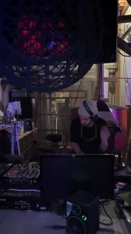 A DJ wearing headphones and a beanie, a black t-shirt and a leather harness over it, is mixing music on a DJ controller. He is standing behind a black table with a computer monitor on it. In front of the table there is a black speaker with a green ring. The room has a rustic aesthetic with wooden beams and panels.  There's a decorative macrame lamp above the DJ with a red light shining through. A person can be seen through a window in the background. There is a wood burning stove with a silver pipe behind the DJ.
