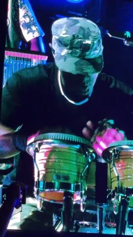 The video shows a concert with multiple screens displaying performances. In the first few seconds, a person wearing a camouflage hat and a black shirt is seen playing bongos. Their hands are blurred as they strike the drums, which are lit with green and pink lights. The stage is dark, with some colorful lighting in the background. The perspective then shifts to a close-up of another performer wearing a green military-style hat, sunglasses, and a patterned shirt. They are singing into a microphone and gesturing with their hands. The crowd is visible in silhouette at the bottom of the screen, holding up phones to record. The final shot is a wider view of the stage, showing the performer playing bongos on a large screen, with bright blue and green lights illuminating the rest of the stage and the text "PARK" visible on a banner.