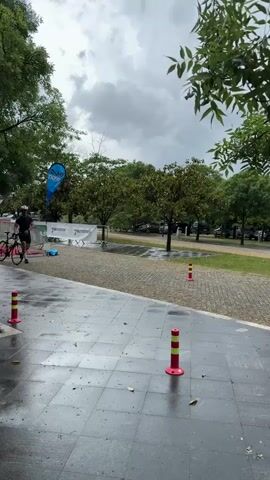 A man wearing a helmet, green short-sleeved shirt and black shorts is shown running with his bicycle. The tile ground is wet. There are orange and yellow bollards. There are green trees in the background. It's cloudy.