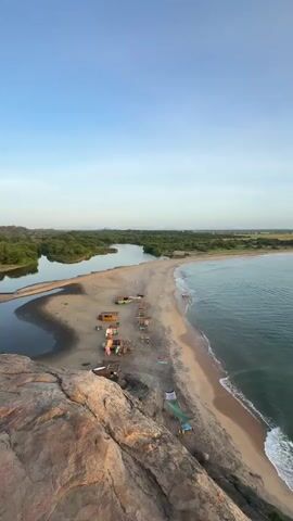 This is a drone video of a scenic coastal landscape in Shastrawela, Sri Lanka. The video begins with an aerial view of a sandy beach, where several small huts or stalls are set up, some with colorful fabrics displayed. A calm body of water, likely a lagoon or river, is visible to the left of the beach, bordered by lush green trees. The ocean stretches out to the right, with gentle waves lapping at the shore.

As the drone ascends and pans, the vast expanse of the turquoise ocean and the curved coastline become more apparent. The sky is a clear, pale blue, transitioning to a warmer hue as the video progresses, suggesting it is either early morning or late afternoon, likely close to sunset given the soft light. The overall mood is peaceful and serene, showcasing the natural beauty of the location.

Further along the coast, rocky outcrops and dense green vegetation are visible, adding to the rugged charm of the landscape. The video captures the interplay of sand, sea, and foliage, highlighting a tranquil beach environment with a sense of remote beauty. There is no visible text in the video.