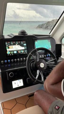 The camera is positioned inside the cabin of a boat, showing the dashboard and a person operating the vessel. The dashboard features two large digital screens displaying navigation information, various control buttons, a steering wheel with the "SHADOWPALM" logo, and a small display showing numerical values. A smartphone is mounted on the right side of the dashboard. The person's hands are visible as they adjust the steering wheel and manipulate switches on the dashboard. Outside the window, the ocean is visible, with a cloudy sky and light rain on the window. The interior of the boat has a beige seat and a patterned brown floor.