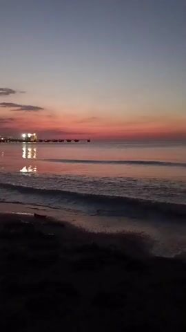 This video captures a serene sunset over the ocean at Jangkar, Indonesia. The scene is a beach at twilight, with the sky transitioning through shades of deep blue, soft purple, and warm pink. The calm sea reflects the fading colors of the sky and the lights of a distant pier, creating a shimmering pathway on the water's surface. Gentle waves lap at the dark, sandy shore, adding to the tranquil atmosphere.

In the foreground, the dark sand and a few scattered pieces of driftwood suggest a natural, undeveloped beach. The pier, visible in the mid-ground, is illuminated with artificial lights, indicating it's a man-made structure, possibly for fishing or leisure. The overall mood is peaceful and contemplative, evoking a sense of calm as day gives way to night. The time of day is clearly sunset, with the sun having already dipped below the horizon. The weather appears calm, with no strong winds or rain evident. The location cues, such as the pier and the specific hues of the sunset, are consistent with a tropical coastal setting like Jangkar, Indonesia. There is no visible text in the video.