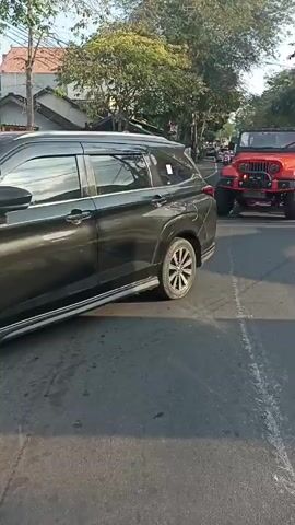 A dark gray SUV is parked on the right side of a paved road, facing away from the camera. An orange Jeep, modified with large off-road tires and a lifted suspension, is driving on the same road, moving towards the camera. The Jeep is in the lane to the left of the SUV. The scene appears to be in a town or city, with trees and buildings lining the street. The sun is shining, casting shadows on the road. The video is shot from a low angle, close to the ground. The focus is on the Jeep as it drives past the parked SUV.