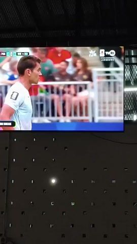 The video shows a large screen displaying a soccer match. A male soccer player wearing a white jersey with the number 5 and a blue trim is the main focus. The score on the screen indicates the match is between two teams, with one team leading 2-0. There is a "HYDRATION BREAK" graphic at the bottom of the screen. The player appears to be in a moment of rest, looking down and to the side. In the background, other players and spectators are visible, along with stadium seating and fencing. The overall environment seems to be an outdoor setting at night, as indicated by the dark background beyond the screen and some faint lighting.
