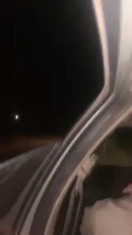 The video shows a night scene viewed through the partially open door of a vehicle. The exterior is dark with sparse lighting from what appear to be streetlights in the distance and oncoming vehicle headlights. The vehicle's interior is visible along the edges of the frame, but details are obscured by the darkness. The camera angle is tilted slightly upwards towards the night sky. The video appears to have been taken while the vehicle was either stopped or moving very slowly.