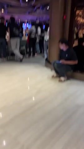The video shows the interior of a casino, filled with people walking around and gambling. The casino has a high ceiling with unusual wooden decorations. The floor is light-colored with a striped pattern. People are playing at various tables and slot machines, most of which are red. The video pans across the room to provide a broad view of the bustling environment. One person is sitting against a pillar, looking at their phone.