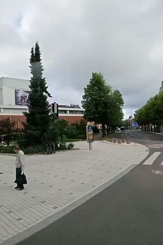 This is a video clip of an outdoor scene in Kuopio, Finland. The scene features a person walking across a paved plaza. The person, who appears to be a man, is wearing a light-colored jacket and dark pants, and is carrying a black bag. Behind the man, there is a large evergreen tree and a modern building with a large poster on its side. The poster shows a black and white image of a person's face with the word "ADAMS" visible in large letters. Beyond the plaza, there is a street lined with trees, and a blue traffic sign with a white arrow pointing left is visible. The weather appears overcast, suggesting it is daytime. The overall atmosphere is calm and ordinary, with no obvious signs of distress or excitement.