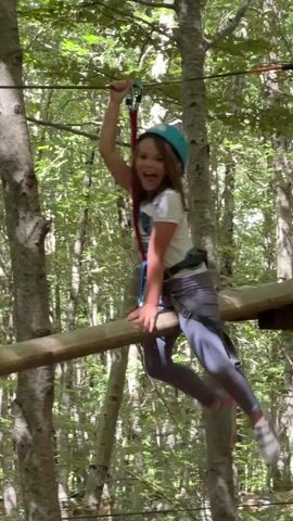 A young girl wearing a blue helmet, white t-shirt, grey pants, and a safety harness hangs from a zip line in a forest. She is smiling and laughing while holding a red and blue zip line handle with her right hand. Her feet dangle near a thick horizontal wooden log, and she occasionally touches it with her shoes. The background consists of numerous trees with green leaves.
