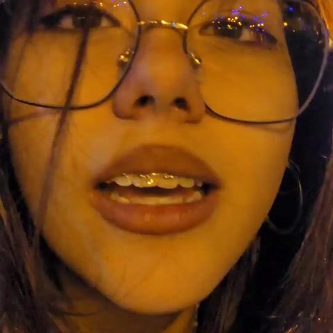 A close-up video features a young woman with brown hair and bangs, wearing metal-framed glasses. She speaks briefly, then rubs her nose with her fingers. The lighting is warm and slightly orange, possibly from street lights. The focus shifts to her hair as she rubs her nose, and the background becomes blurry.