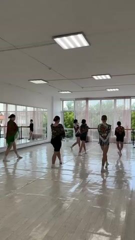A group of seven young female dancers are practicing in a dance studio. The studio has a polished wooden floor, mirrored walls on the left, and large windows with sheer curtains on the right. The room is lit by fluorescent ceiling lights. The dancers, all wearing black dance attire, are spread out across the floor. Some have colorful shawls draped over their shoulders. They are performing a choreographed routine, moving in unison through various poses and formations. The camera is positioned at the front of the room, looking towards the dancers. The scene is calm and focused, indicating a dance class or rehearsal.