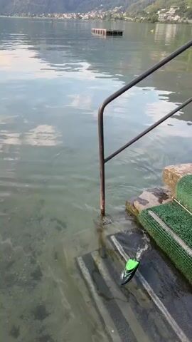 A remote-controlled toy boat with a black hull and a green top is seen moving through clear, shallow water. The boat is leaving a small wake behind it as it navigates over a rocky, gravelly bottom. In the background, there are stone steps leading into the water, and a metal railing for support. The water itself reflects the sky and surrounding landscape, which includes distant mountains and some buildings along the shoreline. There are no people visible in the immediate vicinity of the boat.