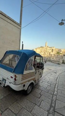 A three-wheeled vehicle with a blue canopy is parked on a cobblestone street. To the left, a silver car is parked. In the distance, a city built into a hillside is visible, with buildings stacked upon each other and a prominent tower. The sky is clear and blue, with some wires crossing overhead. Two people are standing in the distance near the city.