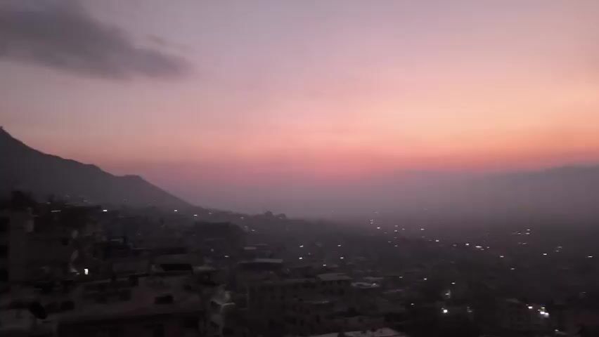 The video shows a cityscape at dusk. The sky is a mix of pink and purple, and there are mountains in the background. The city lights are visible, and the overall scene is somewhat hazy. There is movement in the clouds and lights.
