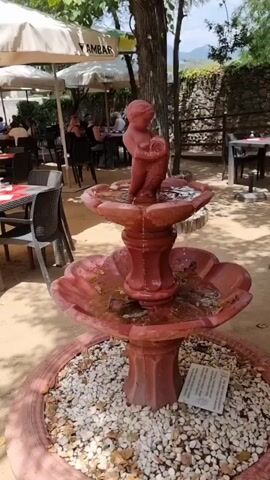 A red three-tiered fountain with a cherub statue on top is in the foreground, surrounded by white pebbles and a few fallen leaves. Water trickles down the cherub and into the tiers of the fountain. Behind the fountain, a restaurant patio is visible with tables, chairs, and umbrellas. People are seated at tables in the background, eating and talking. A stone wall and trees are visible in the far background. The overall scene appears to be a sunny day at an outdoor cafe.