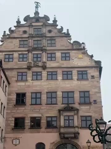 The video shows an exterior view of a historic building, likely a museum, on a cloudy day. The building is made of stone and has multiple floors, with several windows on each floor. It features a decorative facade with a sundial near the top and a small sculpture on the roof. Next to the building is a street with colorful buildings lining it and a decorative lamppost in the foreground. There is a muffled voice in the background, likely from someone speaking.