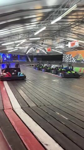 The video shows an indoor go-kart track with several go-karts lined up. One go-kart is in motion on the track. The track is made of wood and has red and white borders. The ceiling is covered with a white tarp and illuminated by fluorescent lights. There are banners and signs visible on the walls, including one that says "KARTING HONDA". The overall environment appears to be an amusement or entertainment venue.