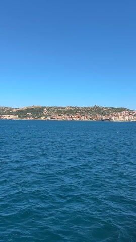 The video opens with a wide shot across a body of water, likely a sea or large lake, under a clear, bright blue sky. In the distance, a coastline is visible, featuring a town built upon a hill. Numerous buildings, predominantly with light-colored walls and some with reddish roofs, are clustered across the hillside, extending along the shore. The water in the foreground is a deep blue, showing ripples and small waves.

As the video progresses, the camera slowly pans to the right. The distant coastline and town remain in view, but gradually, the right side of a vessel, from which the video is being filmed, comes into frame. This reveals a white metal railing running horizontally along the edge of the vessel, and behind it, a grey, somewhat weathered metal door or wall structure. Sunlight casts a distinct shadow diagonally across this grey surface, indicating a bright, sunny day. The panning motion continues, with the distant town receding slightly to the left as more of the boat's structure becomes visible on the right. The water remains calm but rippled throughout the shot.