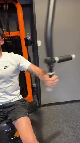 The media file shows a man in athletic wear, a white t-shirt with a Nike logo, and black shorts, exercising on a gym machine. The machine has orange and black components and appears to be a chest press or similar equipment. The man is seated and gripping a black bar with both hands, moving it forward and backward. His arms are visible, and he has a tattoo on his left arm.

The setting is a gym, indicated by the exercise equipment, the rubberized floor, and the wall in the background. The wall is a neutral gray color, and there's a subtle shadow on the floor, suggesting artificial lighting. A small, clear plastic water bottle is visible on top of a part of the orange machinery. A black bag is placed on the seat of the gym equipment.

The activity is clearly a workout. The man's focus is on the exercise. The lighting suggests it could be any time of day, as gyms are typically lit artificially. There are no obvious indications of weather or specific time of day beyond the interior lighting. The man's expression is not clearly visible, but his posture indicates he is engaged in physical exertion.

The visible text is the Nike logo on the man's t-shirt. There is also a white sticker or patch on the gray wall, but the text is not legible. The location is Alès, France, which is not visually indicated within the media itself but provided as context.