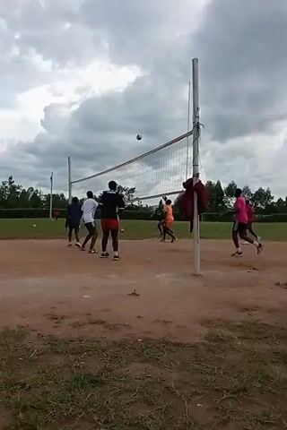 A group of people are playing volleyball on a dirt court under a cloudy sky. There are about 11 people, dressed in various athletic outfits. They are positioned around the volleyball net. The volleyball is in the air and players are either hitting it or preparing to hit it. There are trees visible in the background. A red jacket hangs on one of the volleyball net posts.