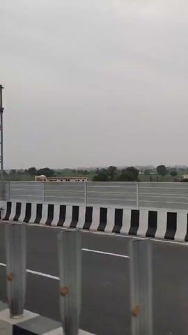 The video shows a drone view moving forward over a highway with a divider. On the left, there is a structure with solar panels. The divider has alternating black and white stripes. There are also traffic signs visible on the road. In the background, there are fields, trees, and buildings. There are also cars on the highway. A cell tower is visible on the right side of the road.
