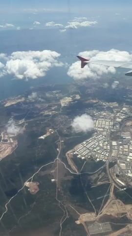 This is a vertical video shot from an airplane window, looking down at a landscape under a clear blue sky with scattered white clouds. The right side of the video shows a portion of an airplane wing with a red wingtip. The land below appears to be a mix of natural terrain, possibly hills or mountains covered in greenery, and a developed area with many buildings, possibly an industrial park or a town, characterized by a grid-like pattern of structures. There are roads and some bodies of water visible in the distance, along with smaller clusters of buildings scattered throughout the landscape. The lighting suggests it is daytime.