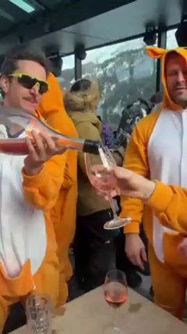 A group of people wearing kangaroo onesies are celebrating at what appears to be a ski lodge. One person pours champagne into a glass. They are at a table with drinks. Other people are milling about the indoor/outdoor space, dressed in various clothing. There is a view of the snowy mountains outside. A large metal structure is visible through the glass wall. People are laughing and socializing. Music is playing.