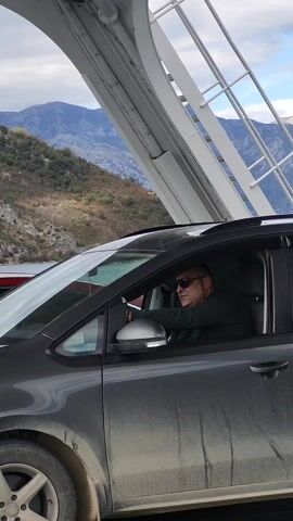 The video shows a man driving a gray car on a ferry. The man is wearing sunglasses and a dark shirt. The car is dirty. In the background, there are mountains and a white metal structure. The sky is cloudy.