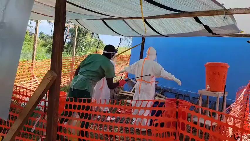 This video captures an outdoor disinfection station in Tchere, Democratic Republic of the Congo. Three individuals are present under a makeshift tent-like structure with a light-colored tarp roof, surrounded by orange plastic safety fencing.

Two individuals are fully clad in white personal protective equipment (PPE) suits, including face shields and gloves, standing with their arms slightly outstretched. A third individual, wearing green scrubs, a white apron, face shield, and gloves, holds a sprayer and is actively spraying one of the PPE-clad individuals, who is standing still with arms open. This suggests a decontamination process.

The scene is set outdoors during daytime, indicated by the natural light. In the background, there is a bright blue wall, lush green foliage, and trees. To the right of the frame, an orange bucket with a spigot rests on a wooden stool. The text "LAVAGE MANO" is partially visible on the bucket, indicating a handwashing station. The overall setup points to a critical point for hygiene and infection control in a field or temporary setting.