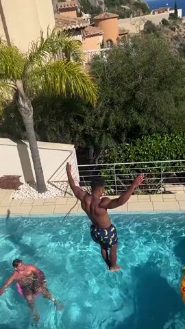 From a high angle, a man jumps into a pool of turquoise water, sending a splash upward. Another man in pink swimwear is already in the water on the left side of the frame, floating on his back. To the right of the jumping man is a pineapple-shaped float with sunglasses. The pool is surrounded by white tiles. Behind the pool is a white railing, lush green foliage, and a palm tree. In the distance, there is a building with an orange roof and a glimpse of the sea.
