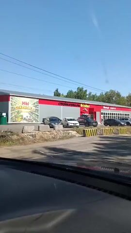 The video shows a car driving past a roadside commercial building. The building has a red and white facade and is identified by signage as "СТРОЙМАТЕРИАЛЫ" (building materials) and "УНИВЕРСАМ" (supermarket). Several cars are parked in front of the building, and one car is entering or exiting the parking lot. The environment is sunny with a clear blue sky. Trees and power lines are visible along the roadside. The video captures a brief view of the street and the surrounding landscape as the vehicle moves forward.