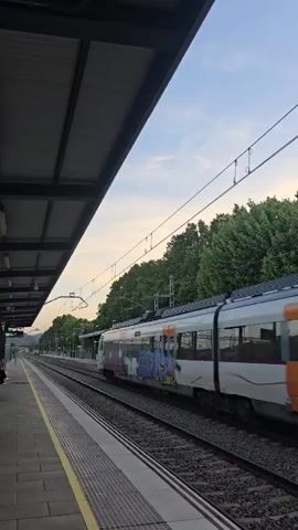 A low-angle shot shows a white and orange Rodalies train, adorned with graffiti on some cars, passing swiftly through a train station at dusk. The train is moving from left to right on the tracks, which run parallel to a yellow-lined concrete platform. The station has a dark metal roof structure visible in the upper left of the frame. Overhead power lines stretch across the sky. In the background, a line of lush green trees borders the tracks, and distant hills are faintly visible under a pale, slightly pink and blue sky, indicating either early morning or late evening. The platform appears empty of people. The scene captures the motion of the train against a serene, natural backdrop. This station is located in la Llagosta, Spain.