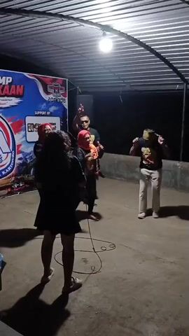 A group of people are dancing and singing in front of a banner. The banner appears to be for an event with the words "JATIM" and "KARTUNURO" visible. There is a microphone and a speaker on the ground. The lighting is dim, with a single bulb overhead. The floor is concrete.