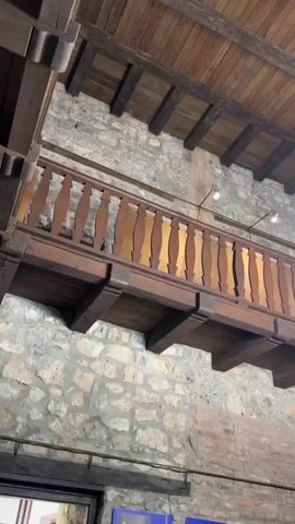 This video shows an interior view of a building with stone walls and a wooden ceiling with exposed beams. A wooden balcony with a wooden railing is visible in the middle of the frame, supported by wooden beams. Two large, ornate chandeliers hang from the ceiling. The left side of the frame shows the balcony and stone wall, while the right side focuses on the chandeliers. The lighting is dim, with bright spotlights attached to the chandeliers and smaller lights on the wall. Some framed items or pictures are visible at the very bottom of the frame. The overall impression is of an old, rustic interior.